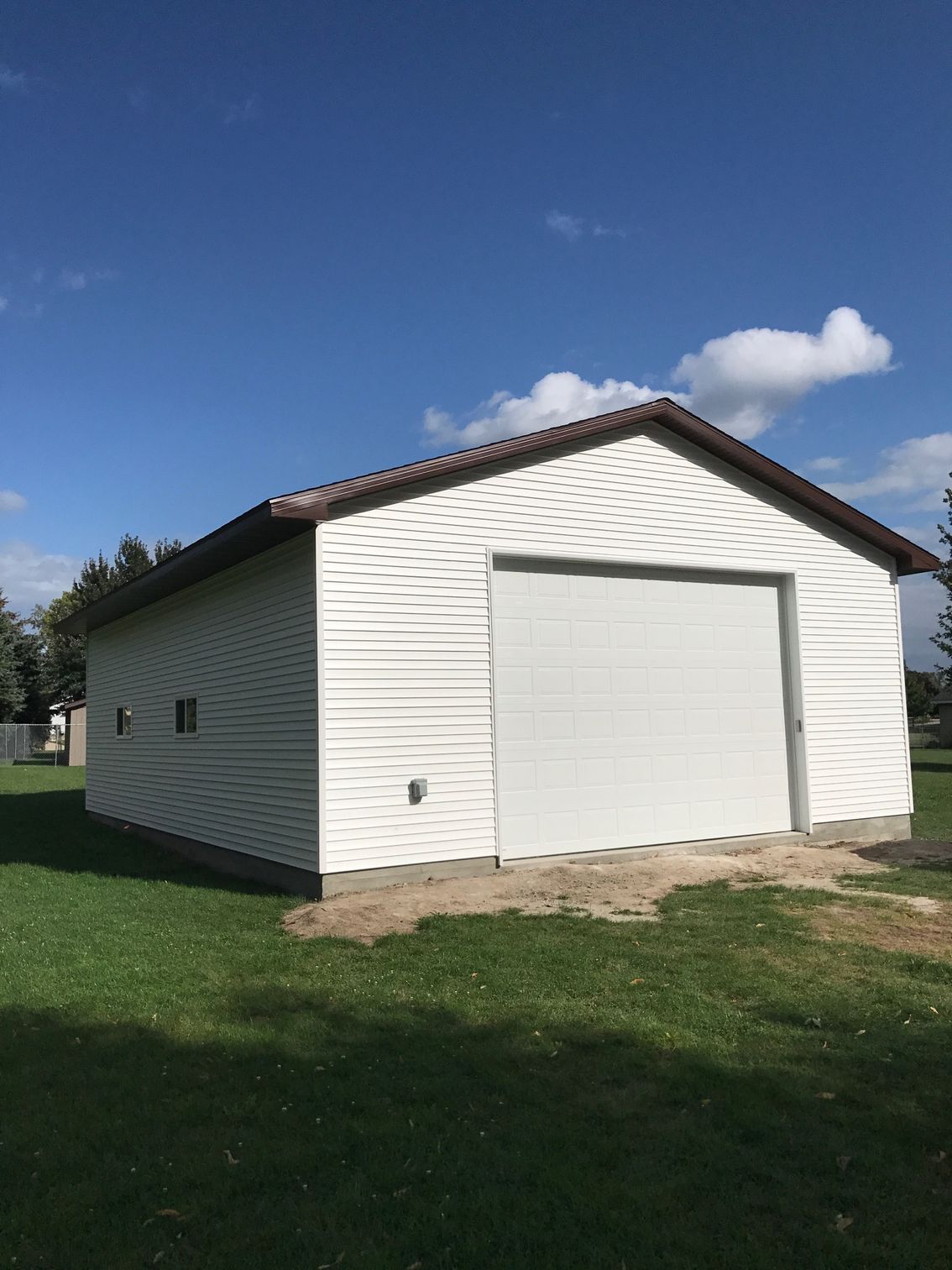White detached garage with a large door on a grassy lot under a blue sky