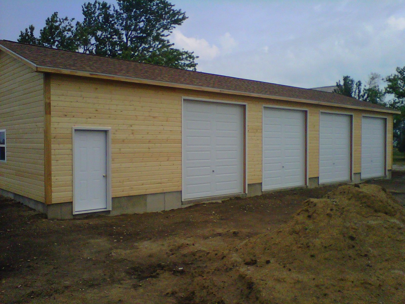 Small wooden building with three white garage doors and one side entry door on a dirt lot.