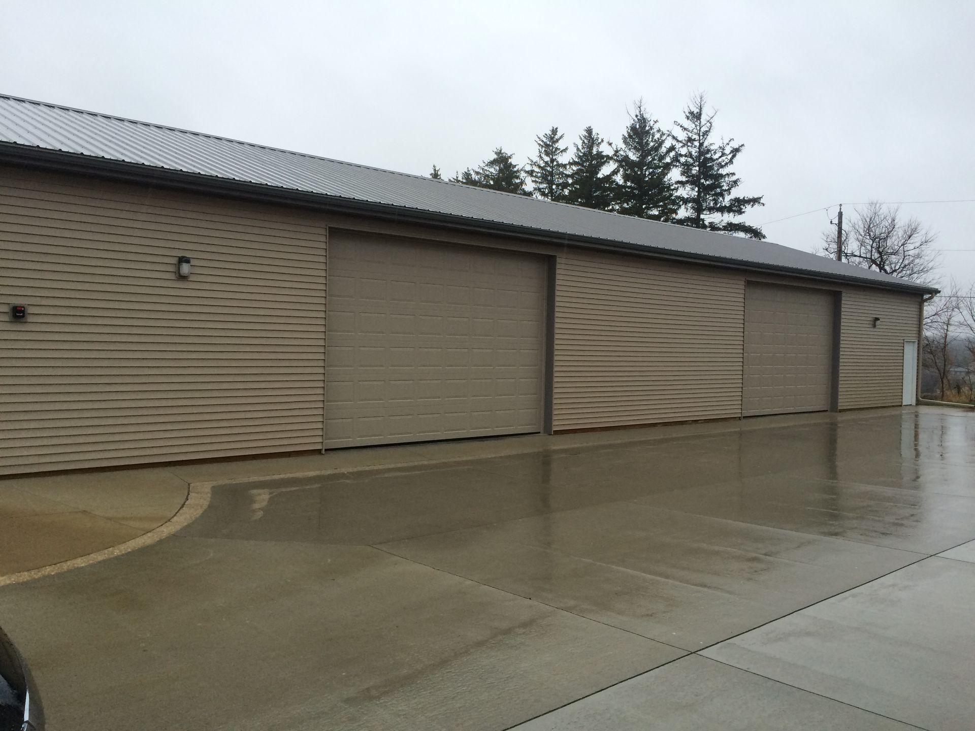 Wet concrete apron outside a beige industrial building with closed garage doors under an overcast sky