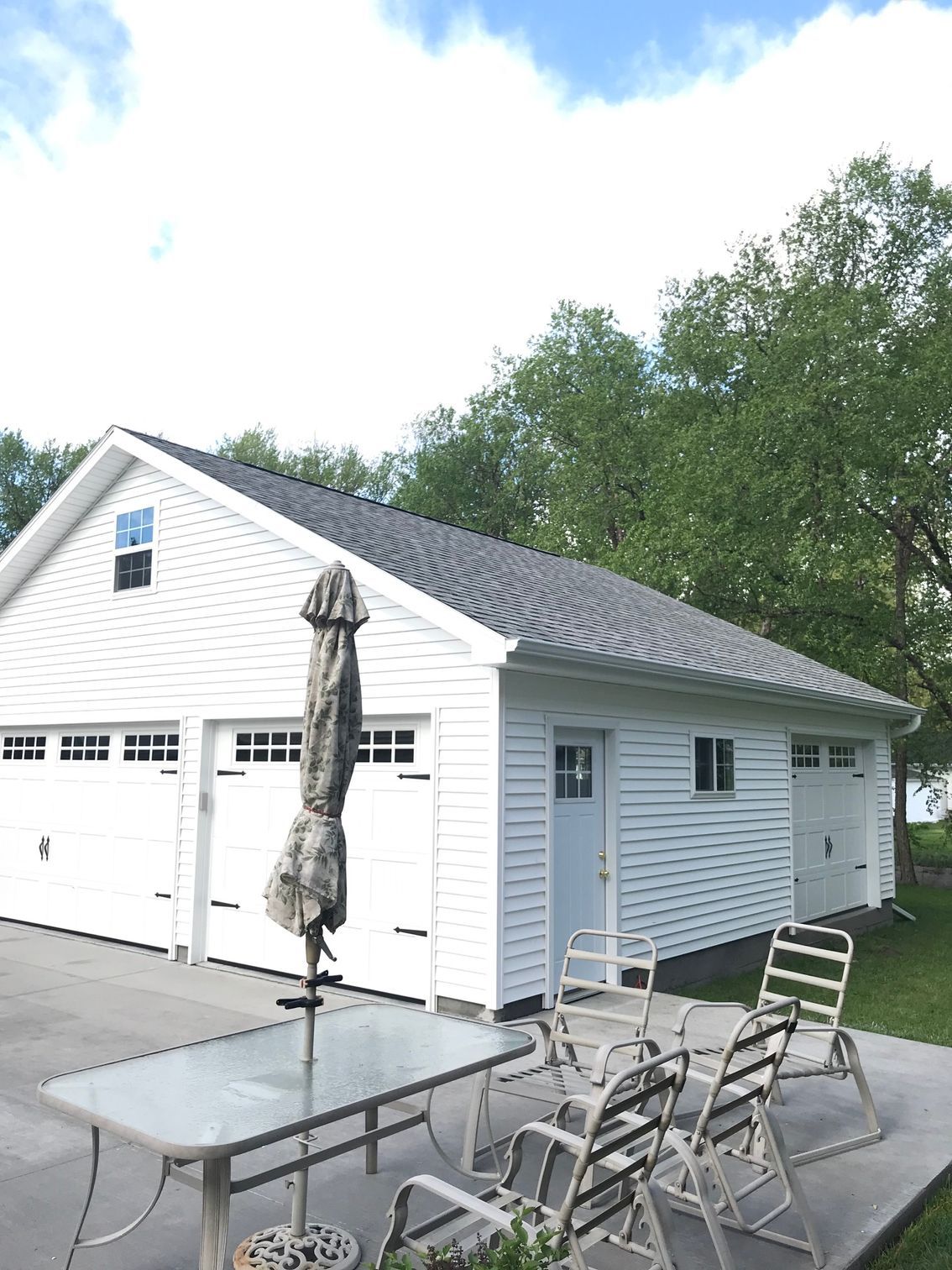 White garage with outdoor patio table, chairs, and closed umbrella beside a driveway.