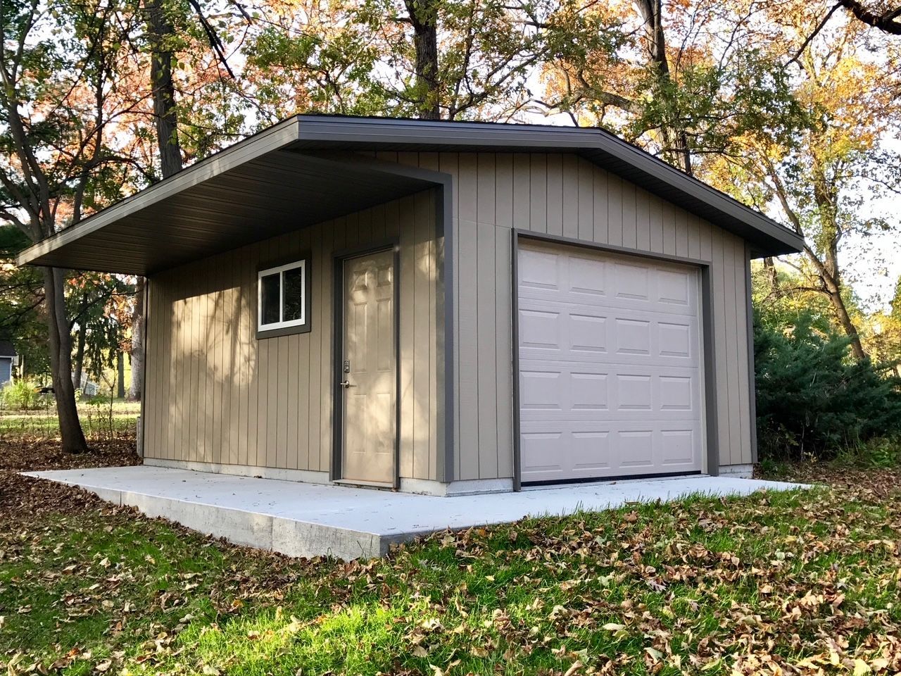 Small beige shed with a gray roof and garage door on a concrete pad in a leafy yard.