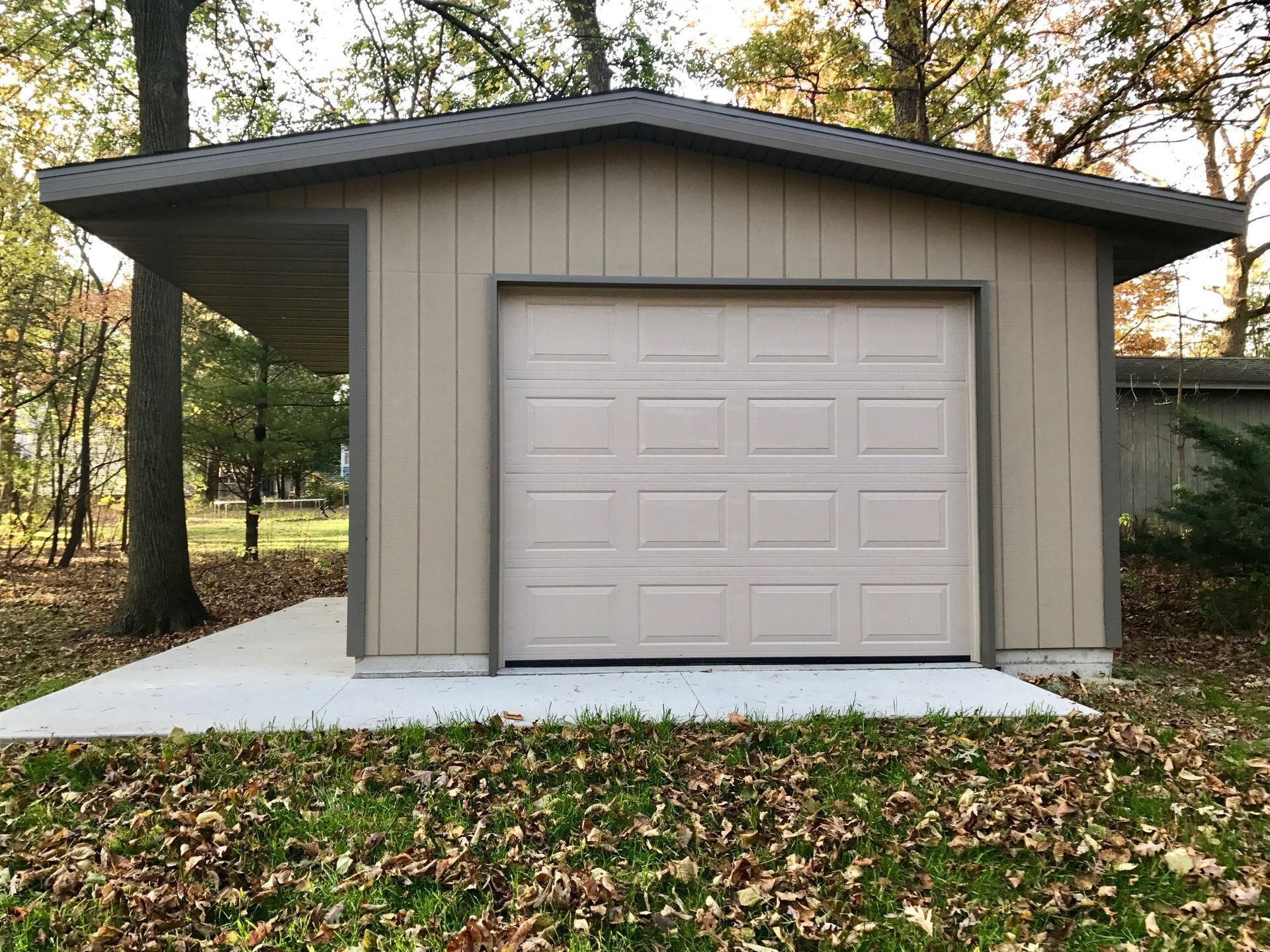 Beige detached garage with a single door, surrounded by trees and fallen leaves