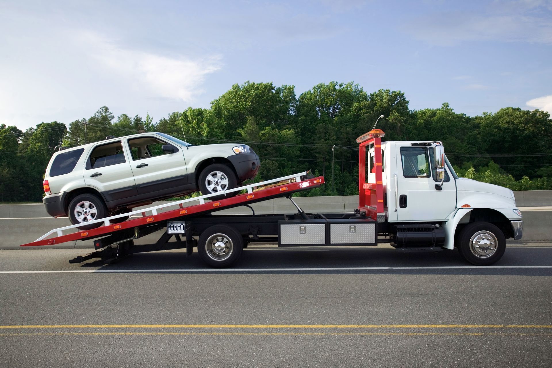 White tow truck carrying a silver SUV on a red flatbed beside a highway.