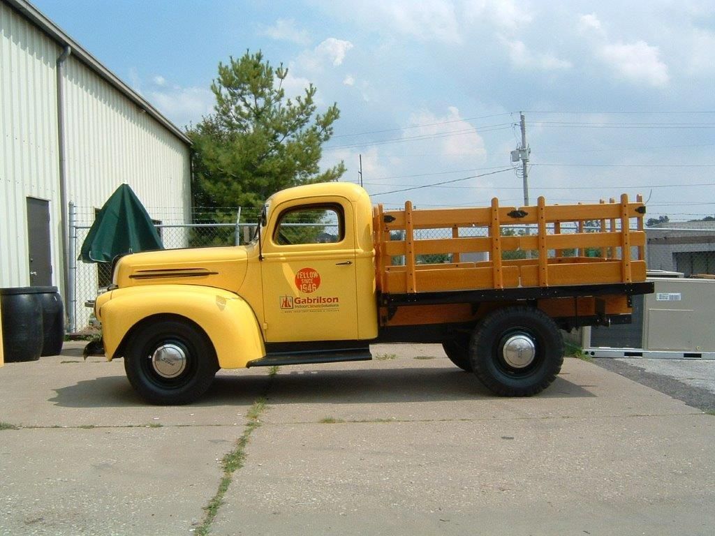 A vintage yellow flatbed truck with a wooden side rack parked on a paved lot beside a building.