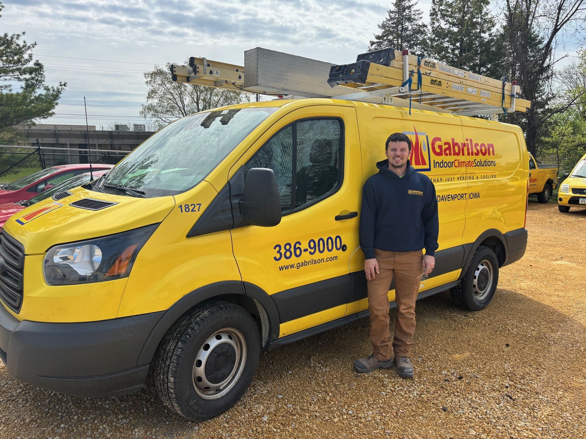 A man stands beside a bright yellow work van with a roof ladder rack and company branding, parked on a gravel lot.