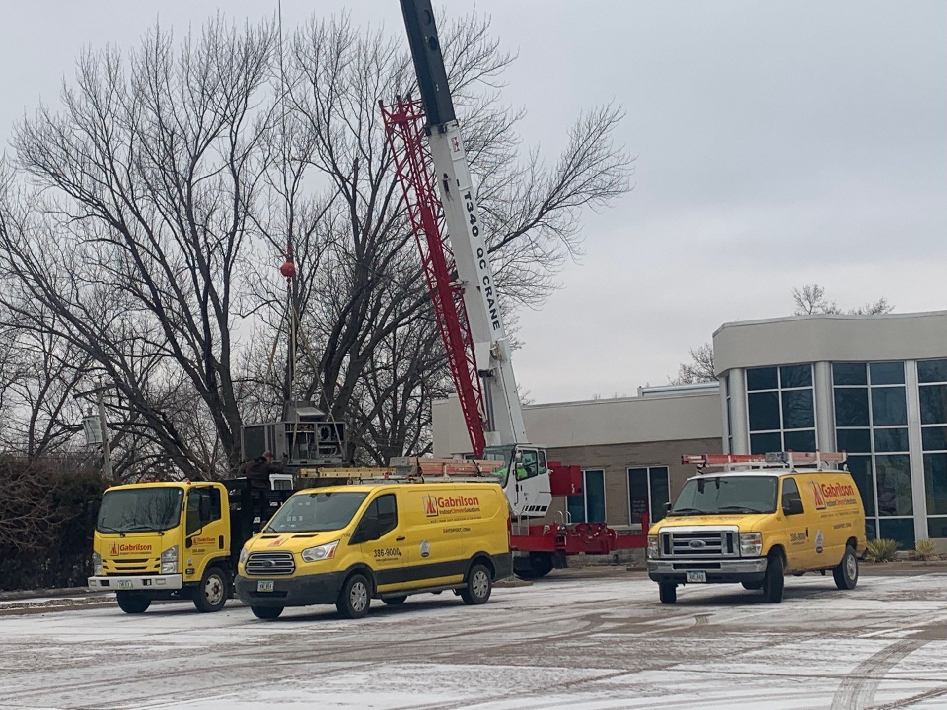 Three yellow utility vehicles and a large mobile crane parked on a snowy, paved lot in front of a building.