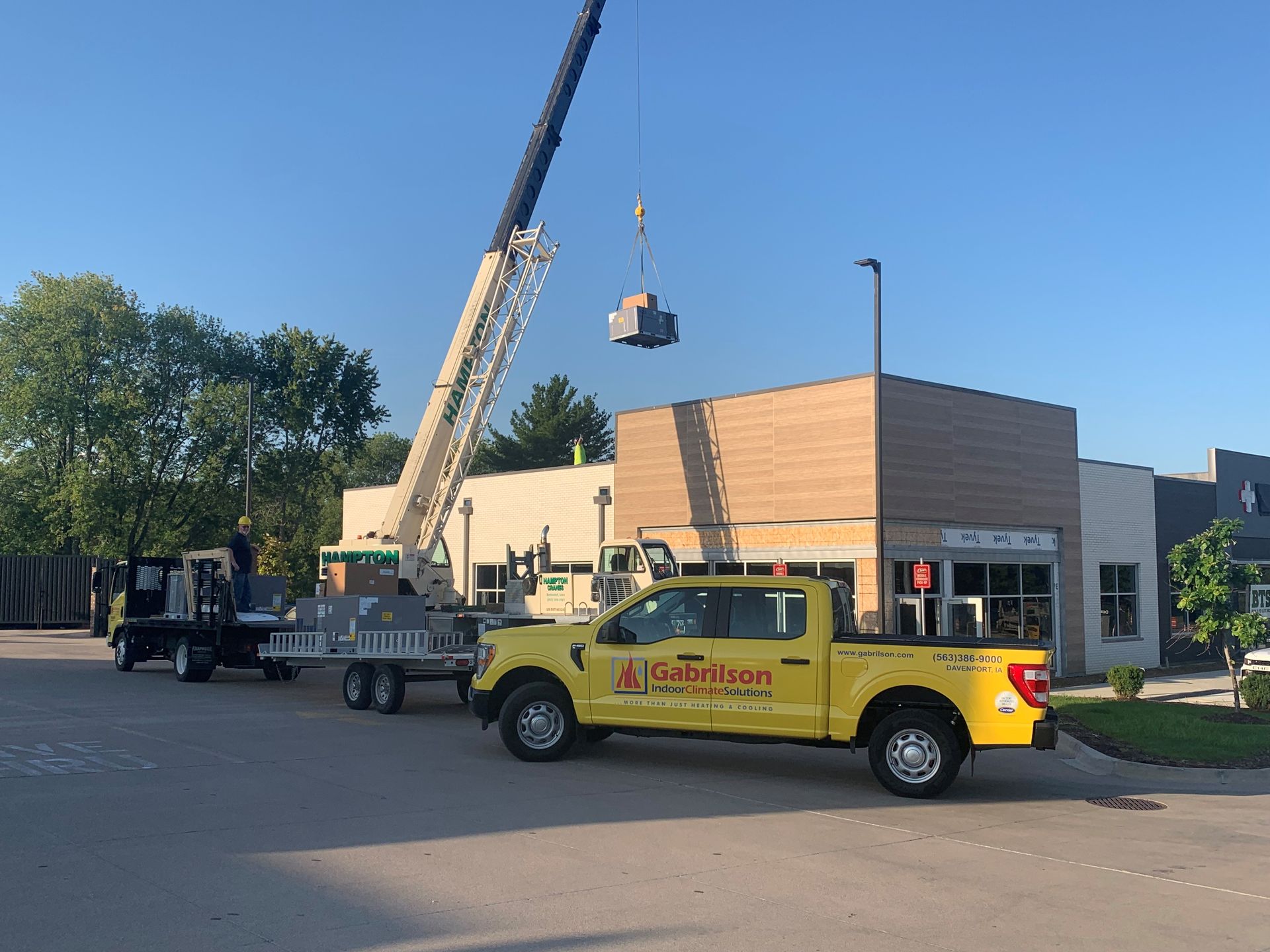 A yellow utility truck sits parked at a construction site where a crane is hoisting equipment onto a building roof.