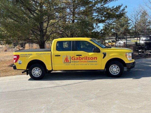 A yellow Gabrilson Indoor Comfort Solutions pickup truck parked on a concrete lot against a backdrop of trees.