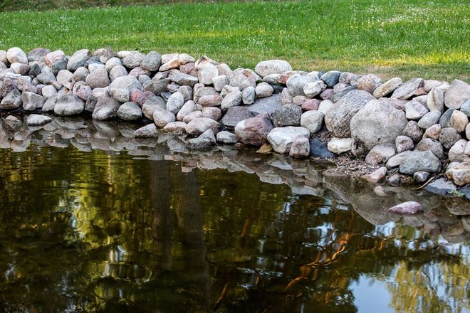 Rock border along a pond reflecting trees and sky, with green grass above the shoreline.