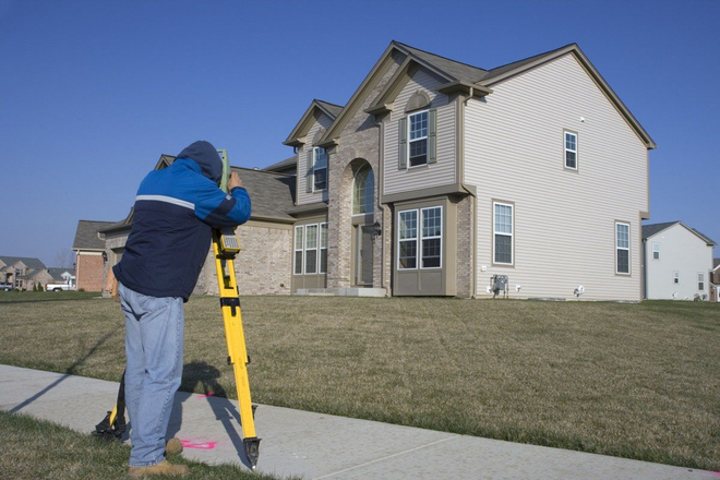 Surveyor using a yellow tripod instrument beside a suburban house on a sunny day