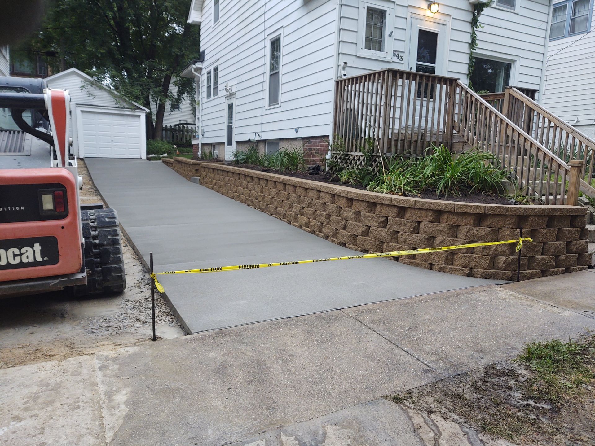 Concrete driveway ramp beside a white house, blocked off with yellow caution tape and a Bobcat machine nearby