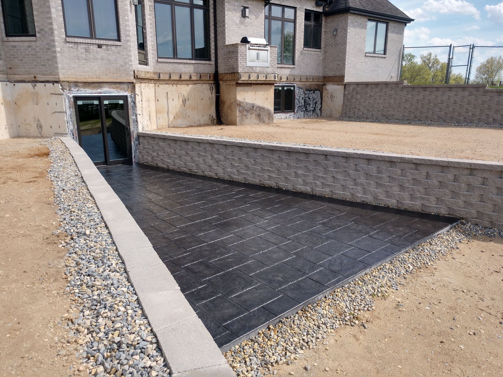 New black patio paving beside a house, bordered by gravel and a low brick retaining wall