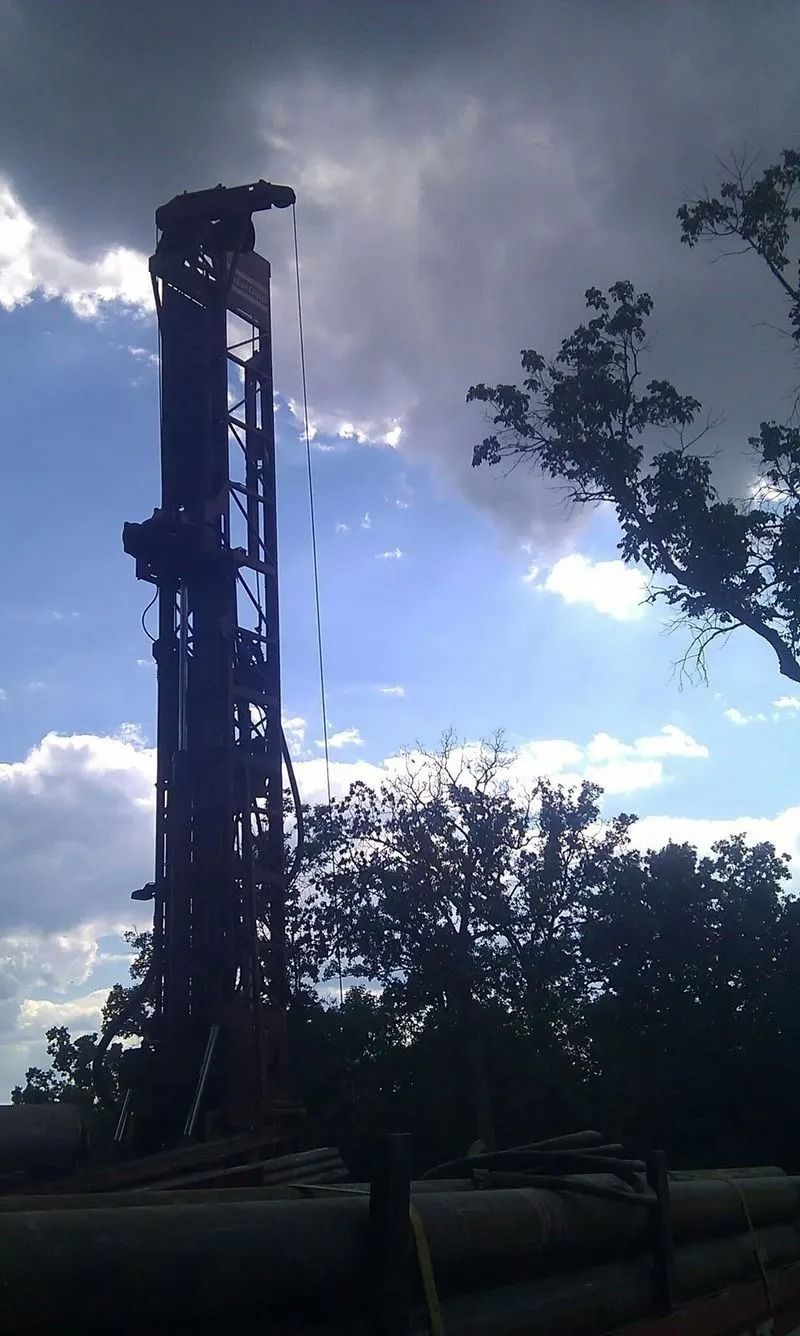 A tall drilling rig against a cloudy sky, near trees and large pipes.