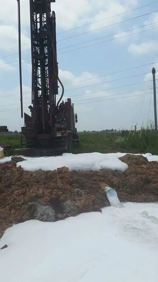 Drilling rig at work, spraying foamy liquid onto surrounding dirt. Blue sky, field background.