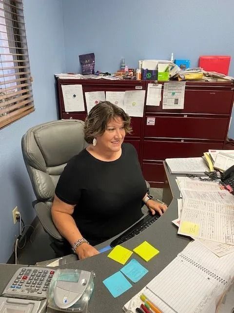 Woman in black shirt sits at a desk with a computer, surrounded by office supplies and papers.