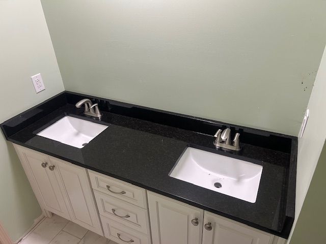 A bathroom vanity with a black countertop, two sinks, and white cabinets. Pale green walls.