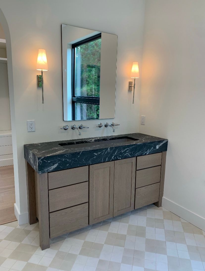 Bathroom vanity with dark countertop, wooden cabinet, and mirror.  Wall sconces flank the mirror. Checkered floor.