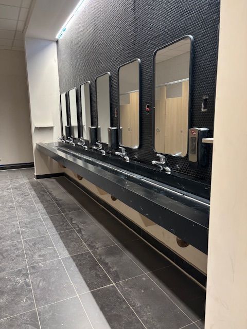 Row of sinks with mirrors in a public restroom. Black countertops and tile floor. Black textured wall.