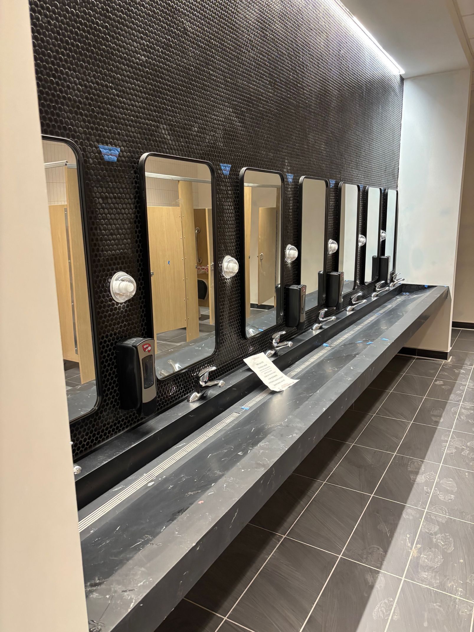 Row of mirrors and sinks in a public restroom with dark tiled wall and neutral colored walls.