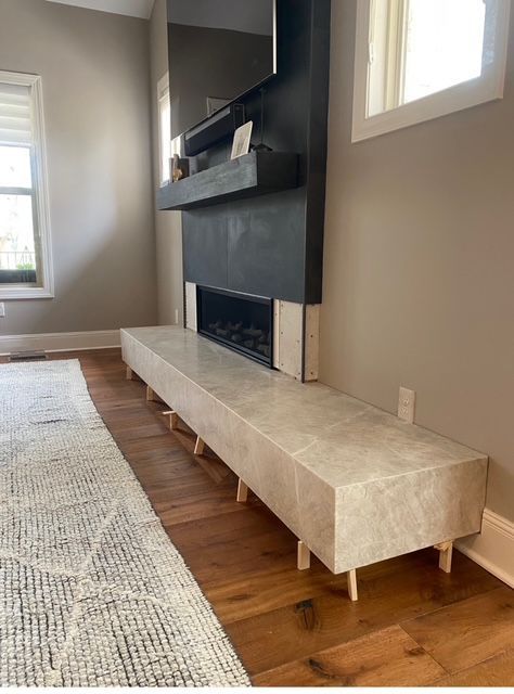 A fireplace with a long, stone-covered bench on wood floors, under a black mantel. A rug lies nearby.
