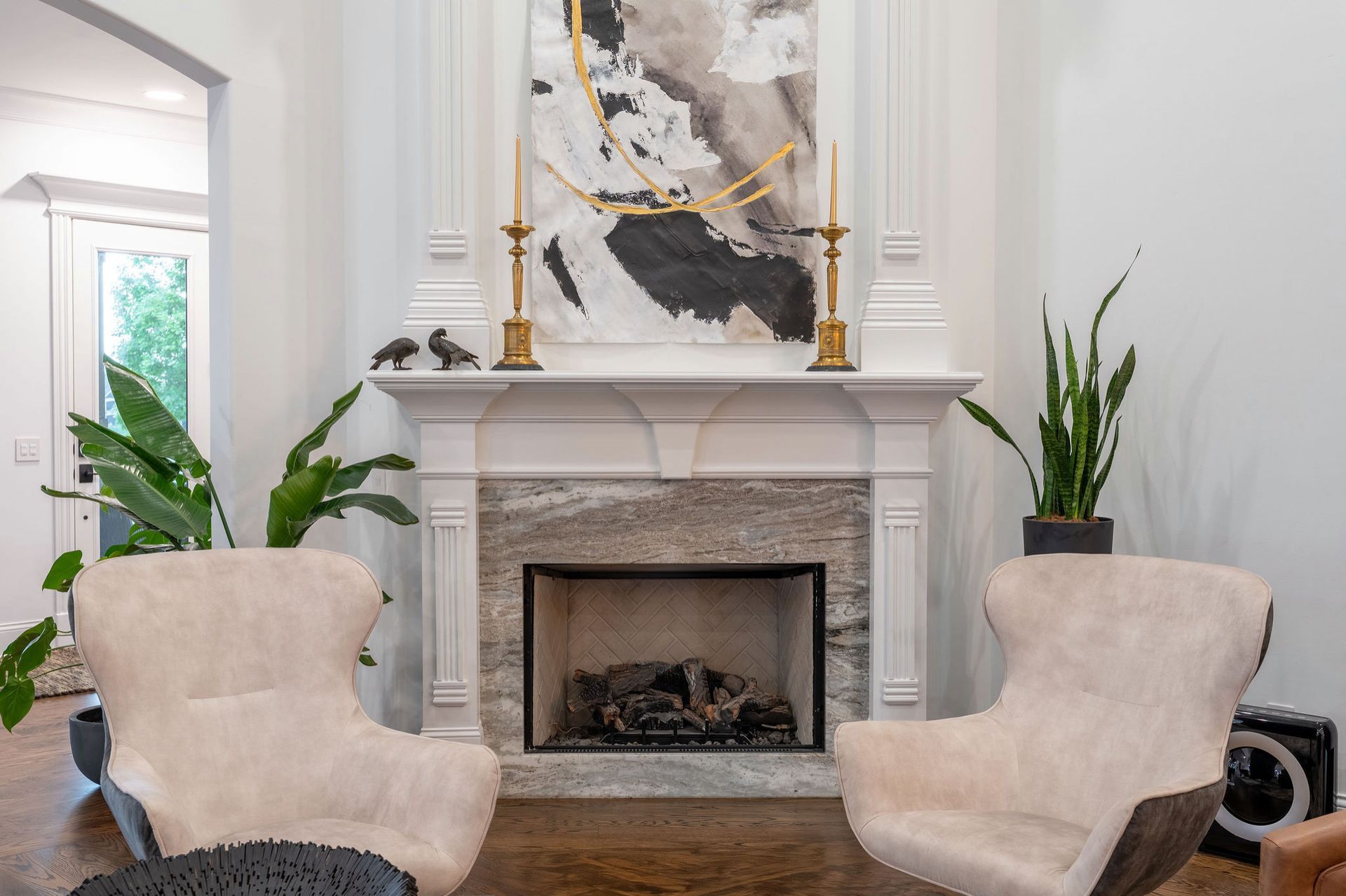 Living room with a fireplace, two white chairs, plants, and abstract artwork.