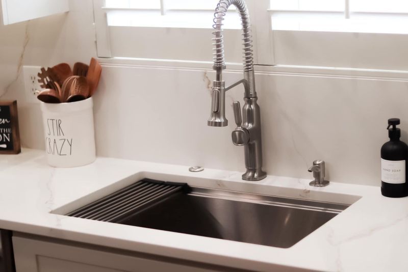 Stainless steel kitchen sink with pull-down faucet, white countertop, and utensils in a jar.