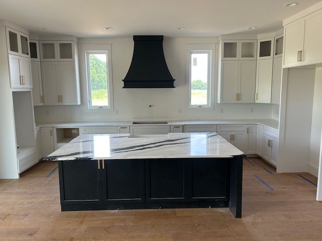 Modern kitchen with white cabinets, black island, range hood. Large marble countertop, windows.