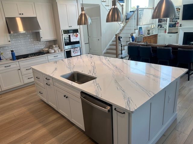 White kitchen island with marble countertop and stainless steel sink and dishwasher; adjacent to a living area.