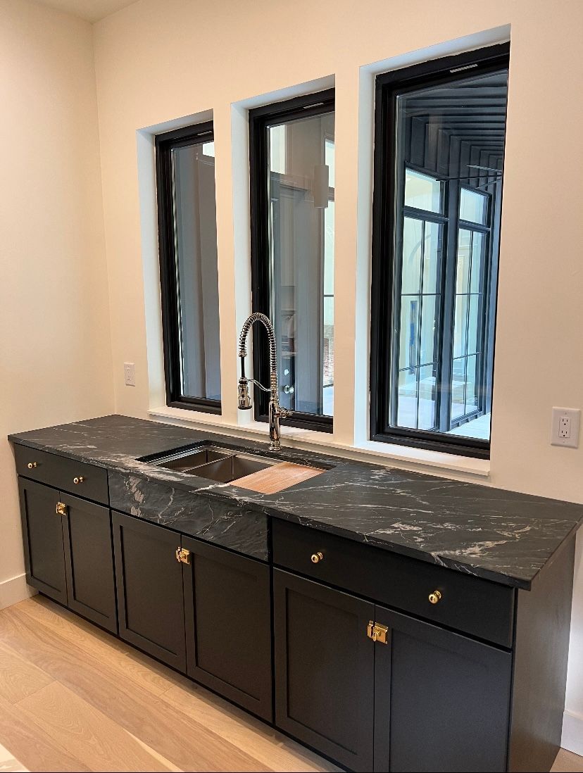 Dark cabinets with a black countertop and a sink beneath three windows.