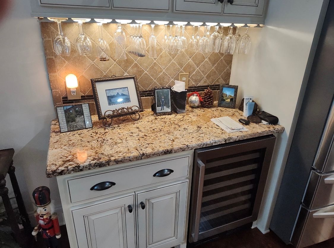 Bar area with a wine fridge, cabinets, and a granite countertop. Wine glasses hang above.