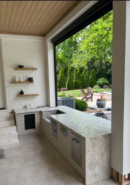 Outdoor kitchen with stone countertop, open to a backyard. Shelves on brick wall.