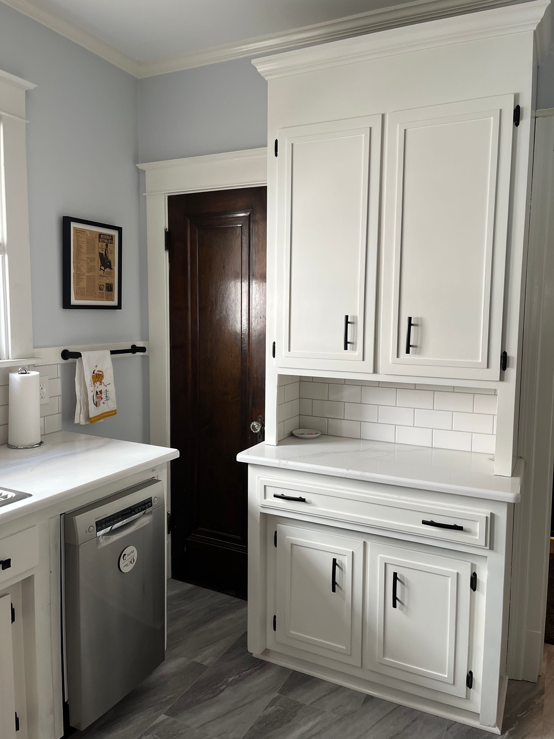 White kitchen cabinets with black handles, dishwasher, and dark brown door. Gray floor, light blue wall. 