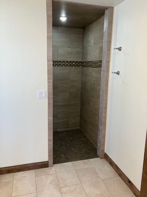 A small walk-in shower with beige tile, bordered with brown trim, set in a room with light-colored walls.