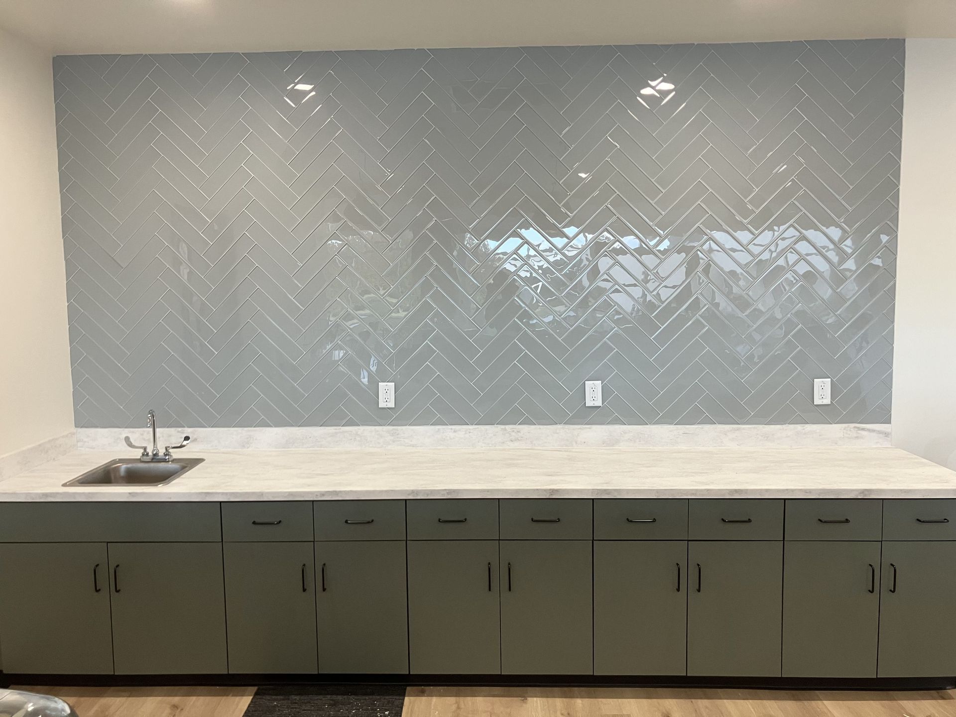 Gray tiled backsplash over gray cabinets with a sink and countertop.
