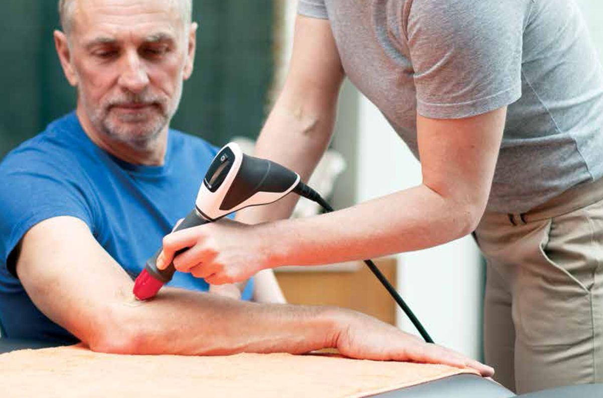 Therapist using a medical device on a patient's arm; both are indoors.