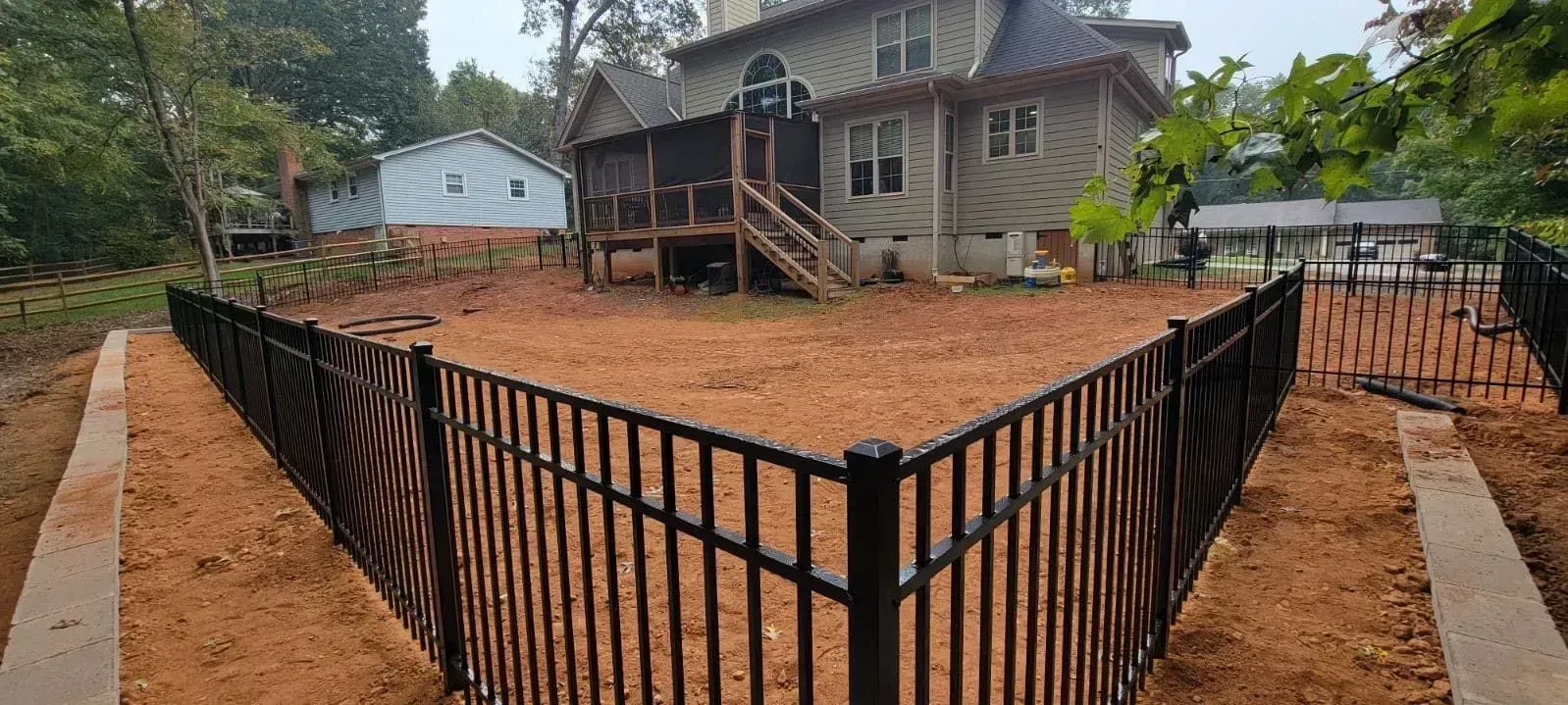 A black metal fence surrounds a yard with a house in the background. The yard is filled with reddish dirt.