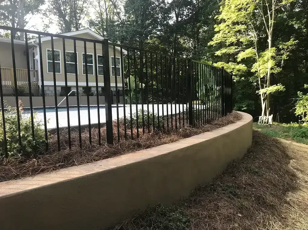 Black metal fence atop a low, curved retaining wall; trees and house in the background.