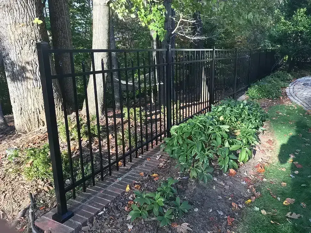 Black metal fence along a brick and earth garden bed with bushes and trees in the background.
