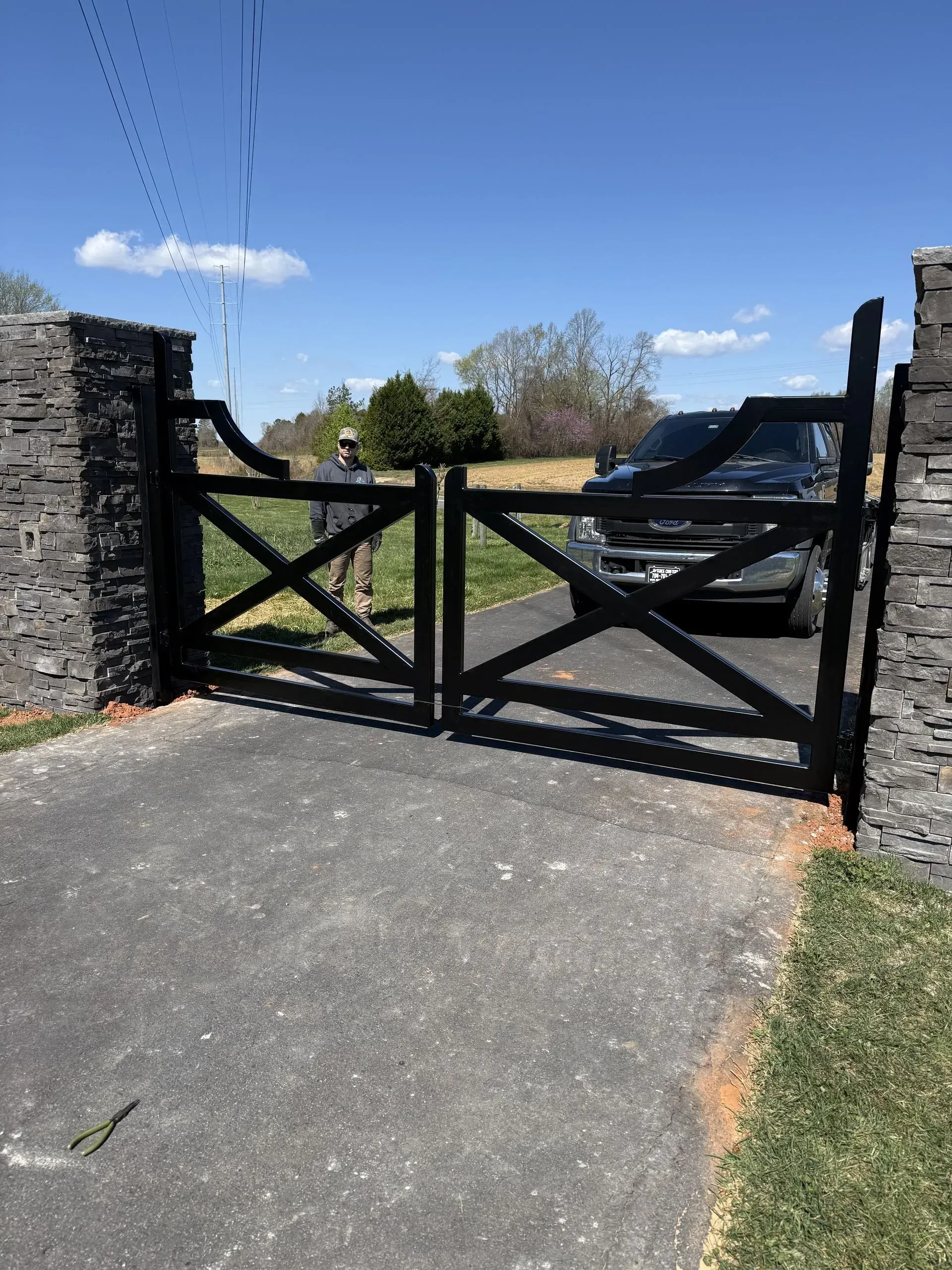 Black wooden gate between stone pillars, with a driveway and a vehicle in the background on a sunny day.