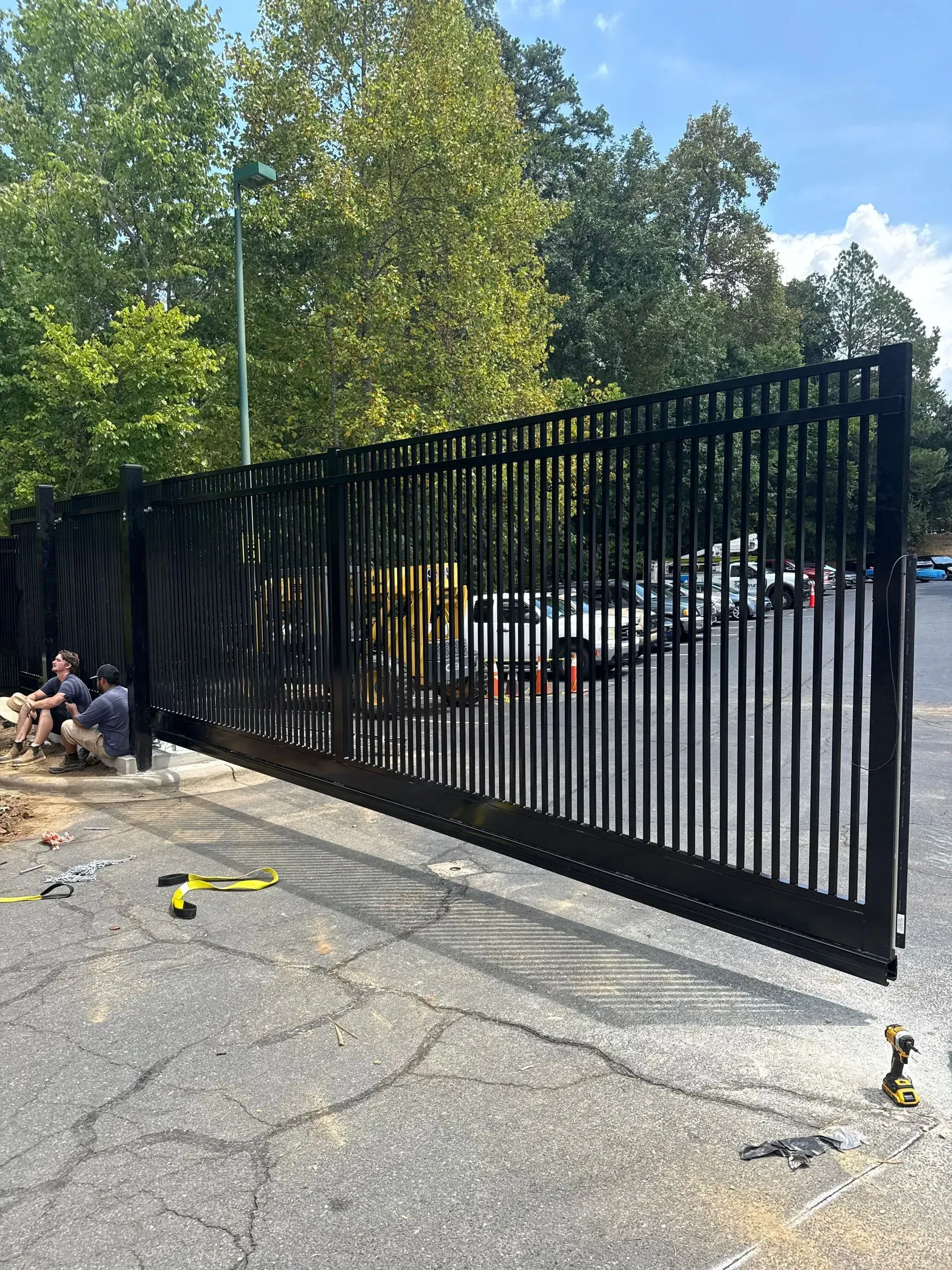 Black metal sliding gate being installed on asphalt with two workers nearby. Trees in background.