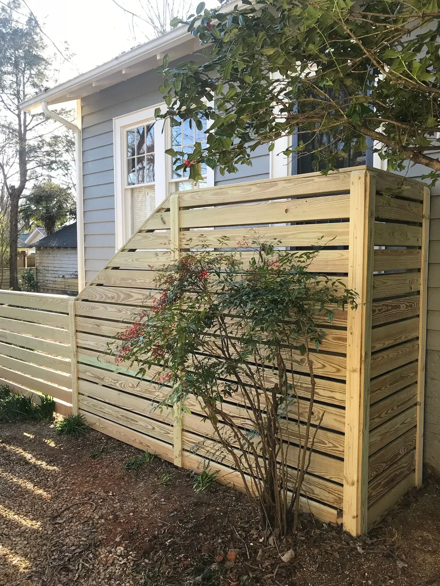 Wooden fence enclosure next to a light blue house, with a small bush growing inside the fence.