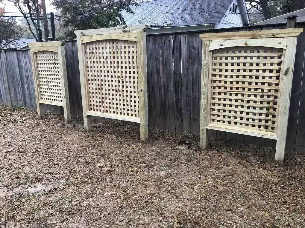 Three wooden lattice panels stand in front of a weathered wooden fence, covered in fallen leaves.
