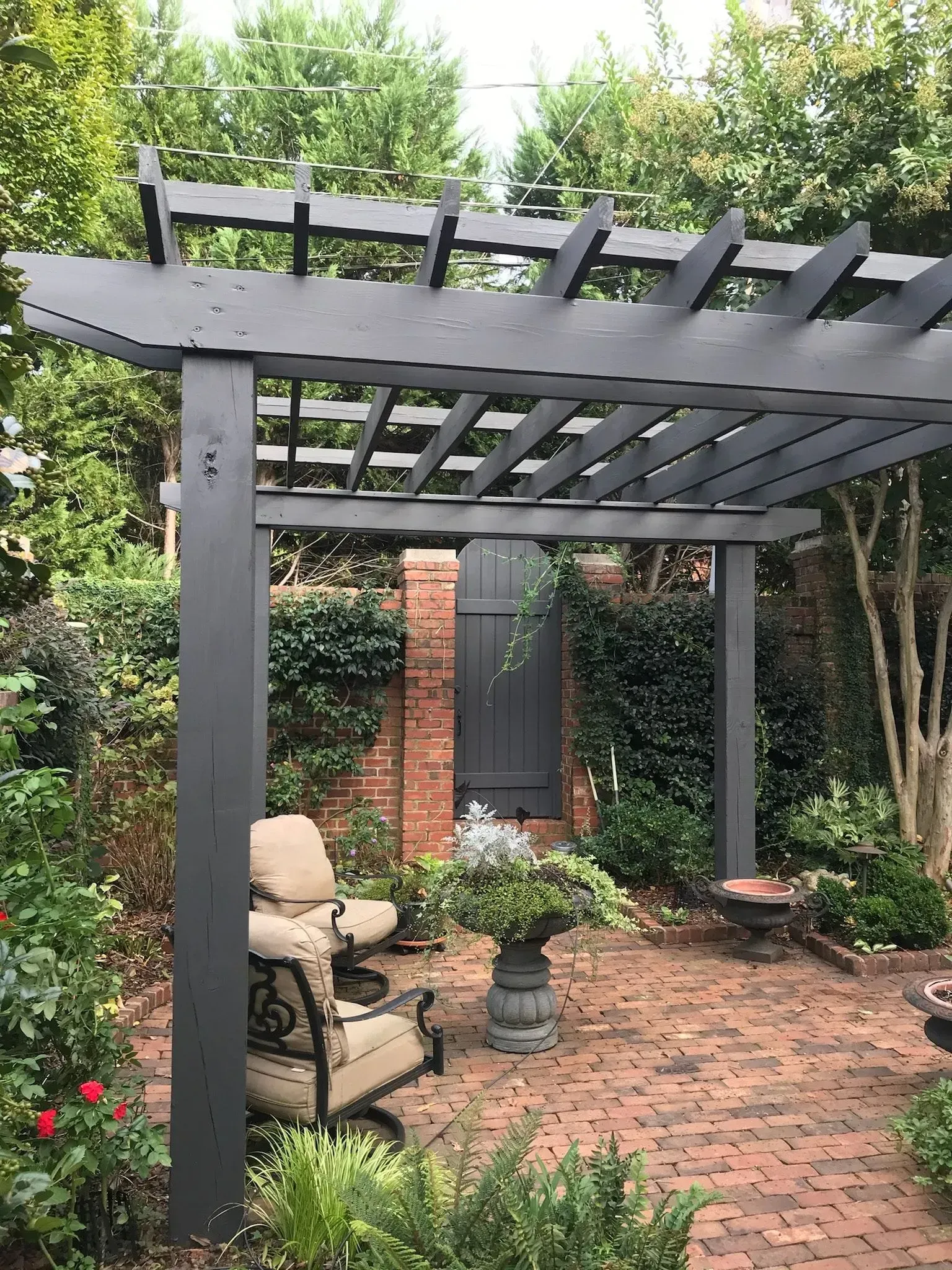 Gray pergola over a brick patio with an armchair, surrounded by plants and brick wall.