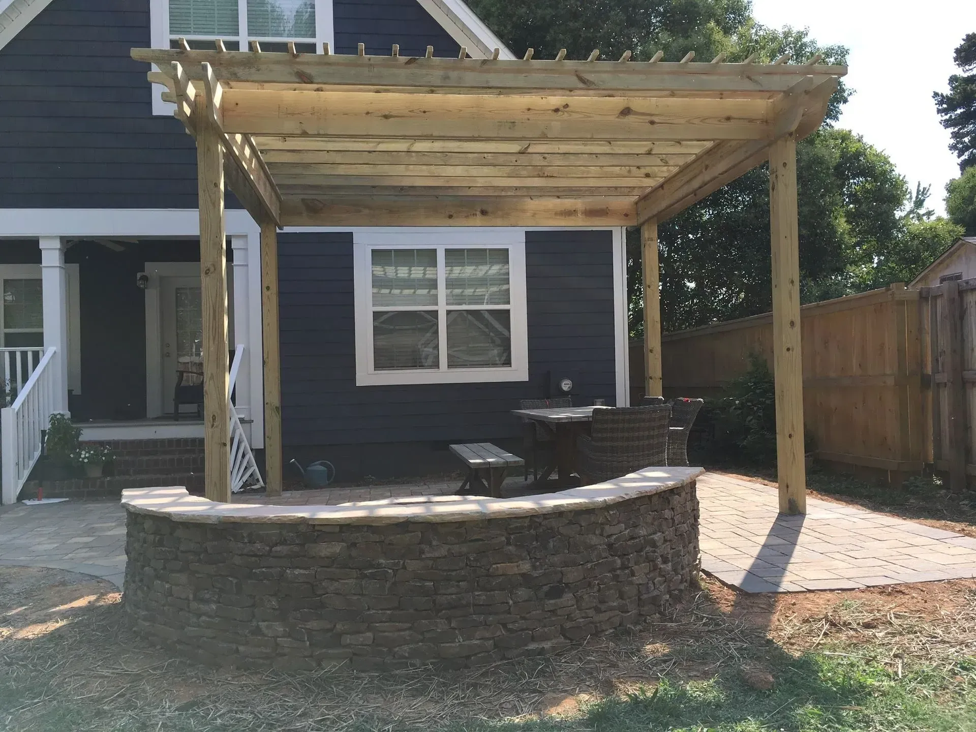Wooden pergola over a stone-walled patio with furniture against a dark blue house.