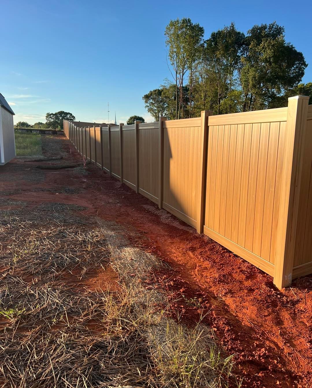 Brown vinyl fence stretches across a red-dirt yard on a sunny day.