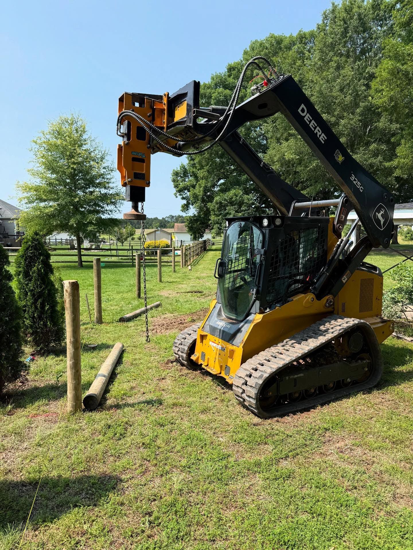 Yellow John Deere skid steer installing fence posts in a grassy yard under a blue sky.