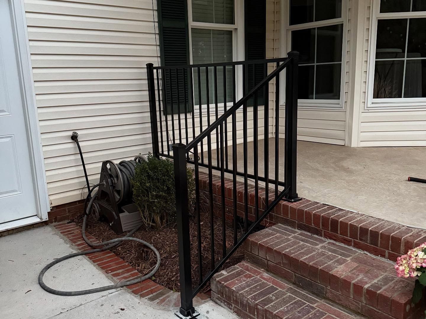 Black metal railing on brick steps of a house with white siding and window. A hose and bush are visible.