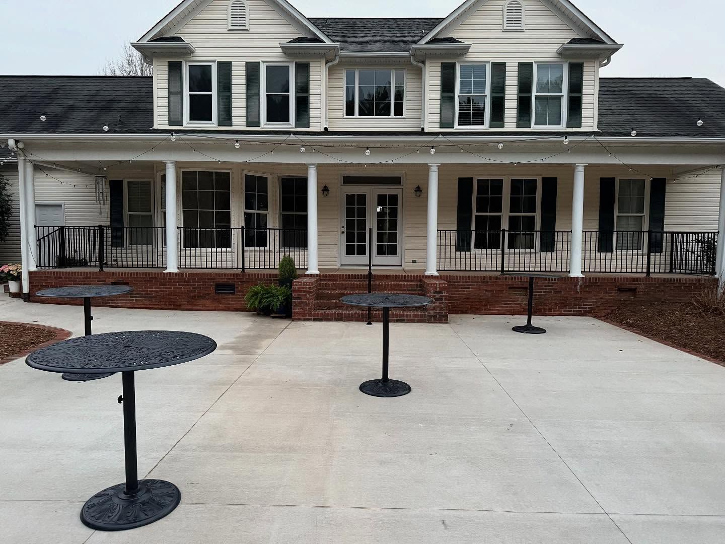 Exterior of a two-story beige house with a porch and black high-top tables on the concrete patio.