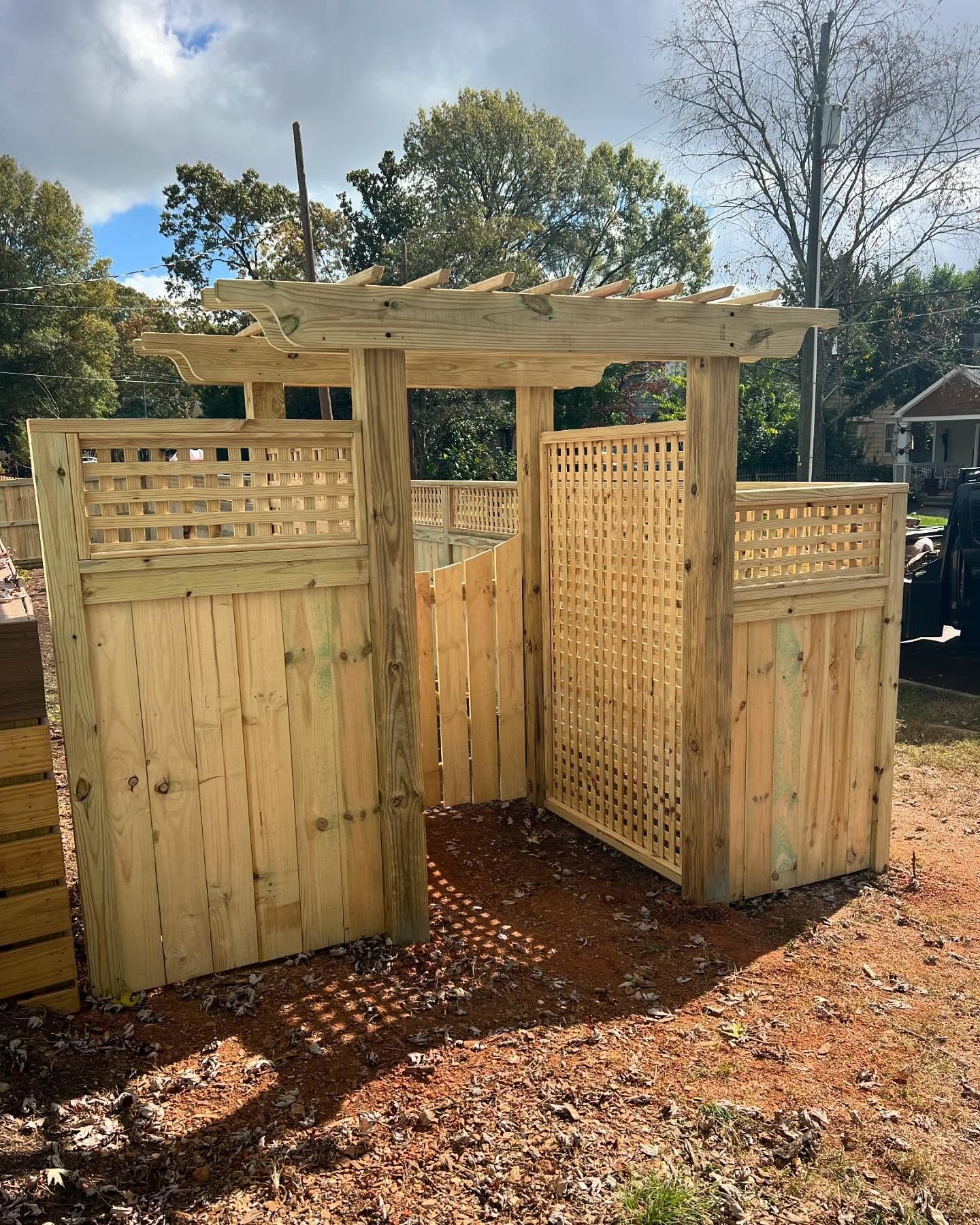 Wooden arbor with trellis panels, entryway to a fenced area in a yard; sunny day.