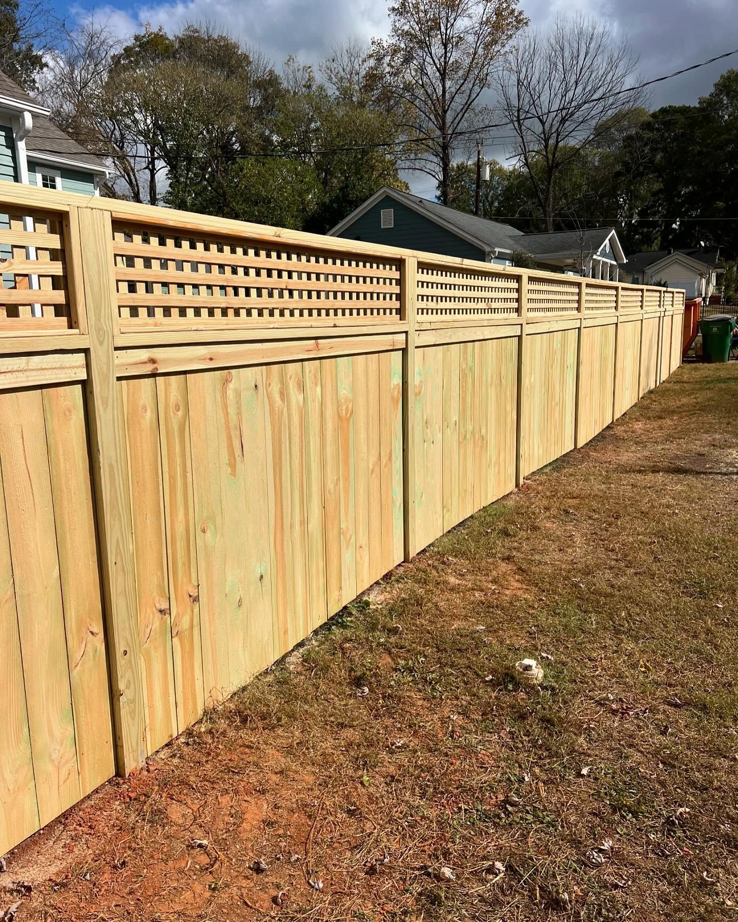 Wooden fence with lattice top, surrounding a grassy area near houses and trees on a cloudy day.
